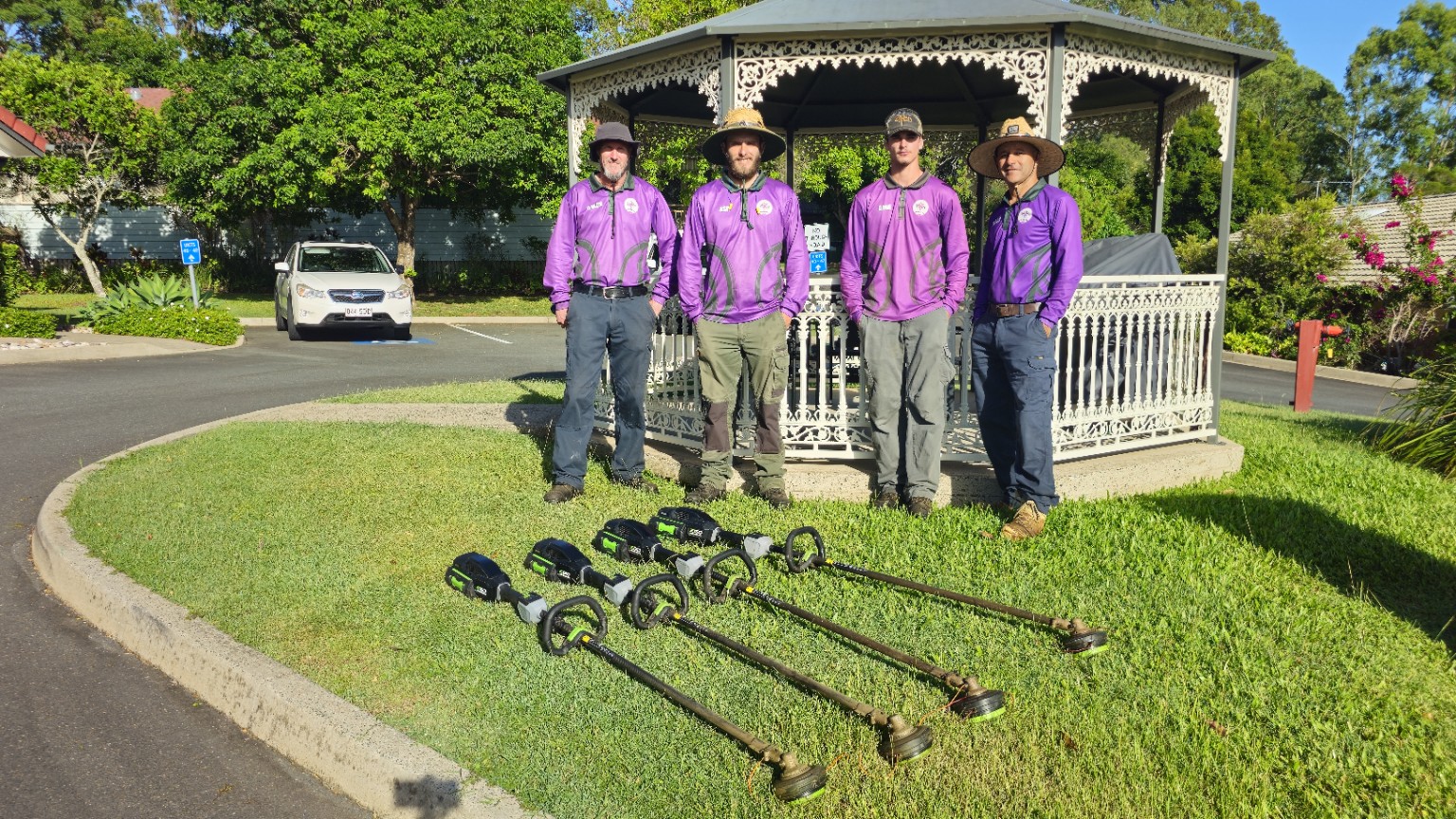 Four Excel Grounds Maintenance team members beside a village gazebo, with battery-powered grounds equipment on the lawn